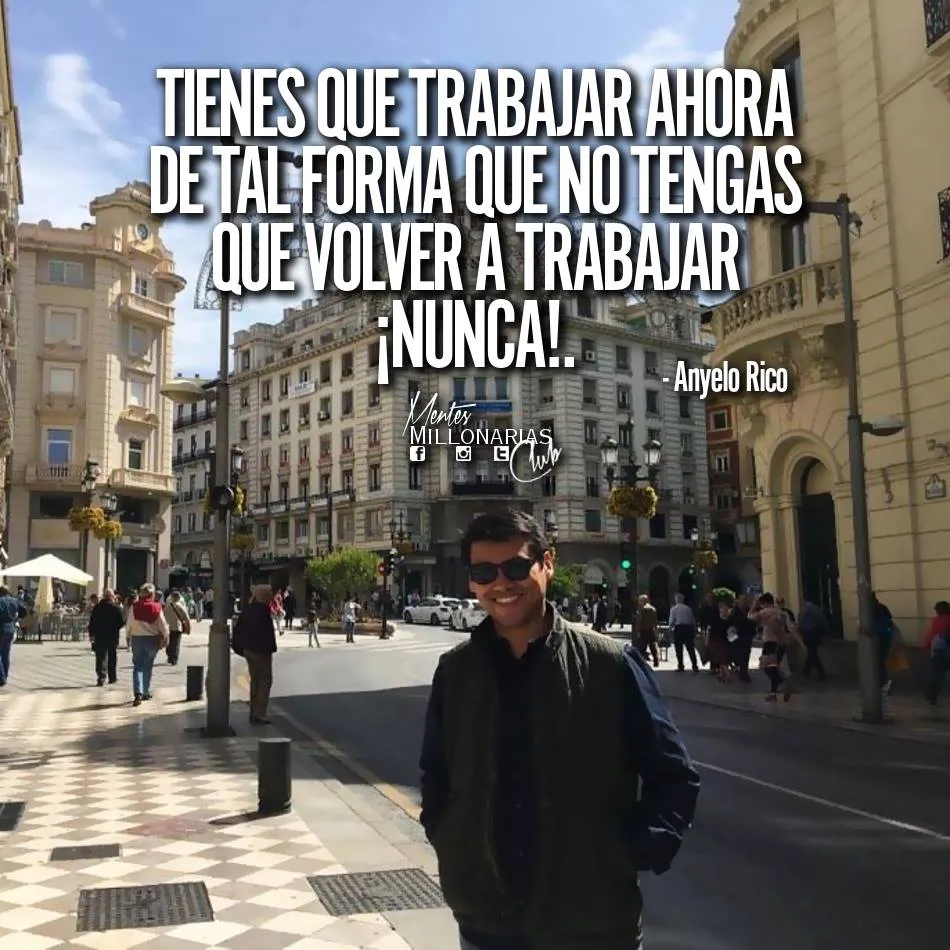 Se ve a un hombre de pie en una calle adoquinada en España. Lleva una chaqueta negra y gafas de sol. La calle está llena de gente caminando. Hay edificios a ambos lados de la calle. El cielo está soleado.